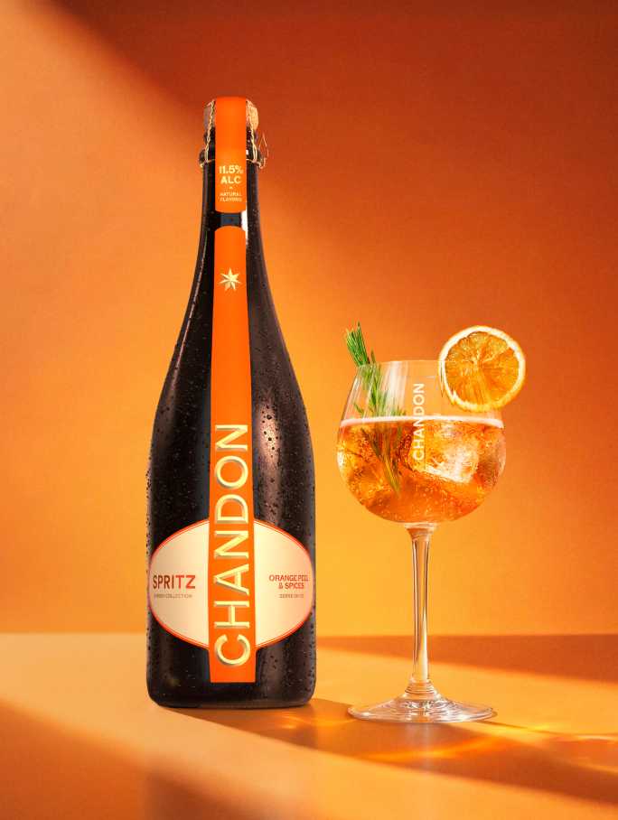 Bouteille de Chandon Spritz, tranche d'orange séchée, brin de romarin et glaçons disposés sur un fond orange à côté du contour embossé d'un verre à vin.
