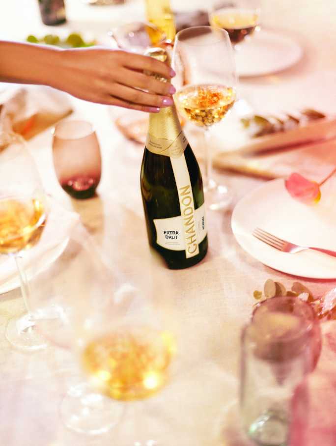 Mano femenina descorchando una botella de CHANDON Extra Brut en una mesa elegante con copas de vino.