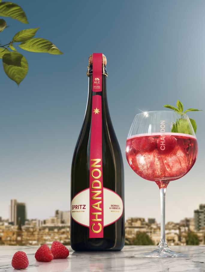 A bottle of Chandon Spritz Berries & Hibiscus next to a glass of Chandon Spritz cocktail garnished with fresh raspberries and mint leaves, set against a bright cityscape background under a clear sky.