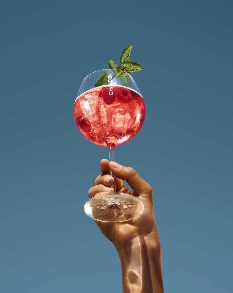 A hand holding up a large wine glass filled with a red Chandon cocktail, garnished with fresh raspberries and mint leaves, set against a clear blue sky.