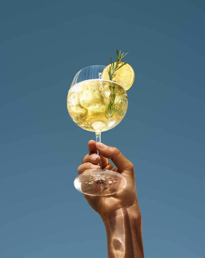 A hand holding up a large wine glass filled with a Chandon cocktail, garnished with a lemon slice and a sprig of rosemary, set against a clear blue sky.