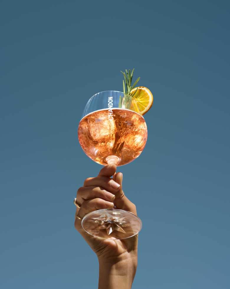 Eine Hand hält ein großes Weinglas, gefüllt mit einem Chandon Spritz Orange Peels & Spices, garniert mit einer getrockneten Orangenscheibe und einem Rosmarinzweig, vor einem klaren blauen Himmel.