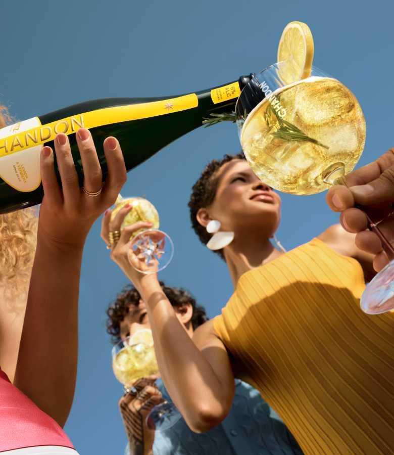  woman in a yellow top and large white earrings smiling upwards as a bottle of Chandon Spritz Lemon & Verbena is poured into a glass in the foreground, with another person holding a glass visible in the background, all under a clear blue sky.