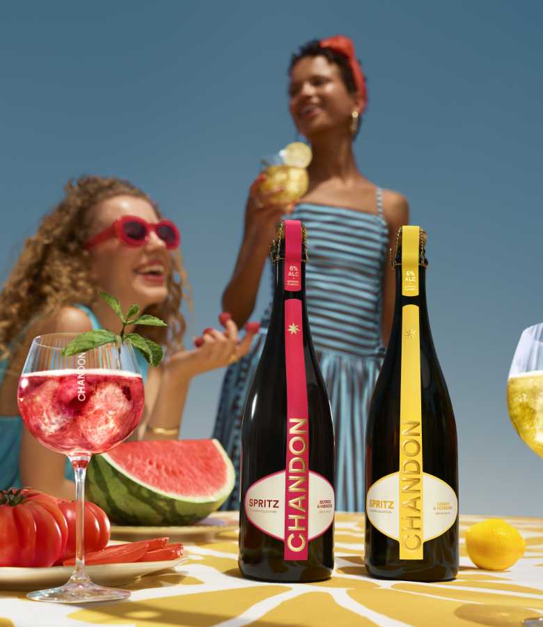 Two bottles of Chandon Spritz (Berries & Hibiscus and Lemon & Verbena) standing on a table with a glass of red Chandon Spritz cocktail, a watermelon slice, and fresh berries. Two smiling women are visible in the blurred background under a clear blue sky.