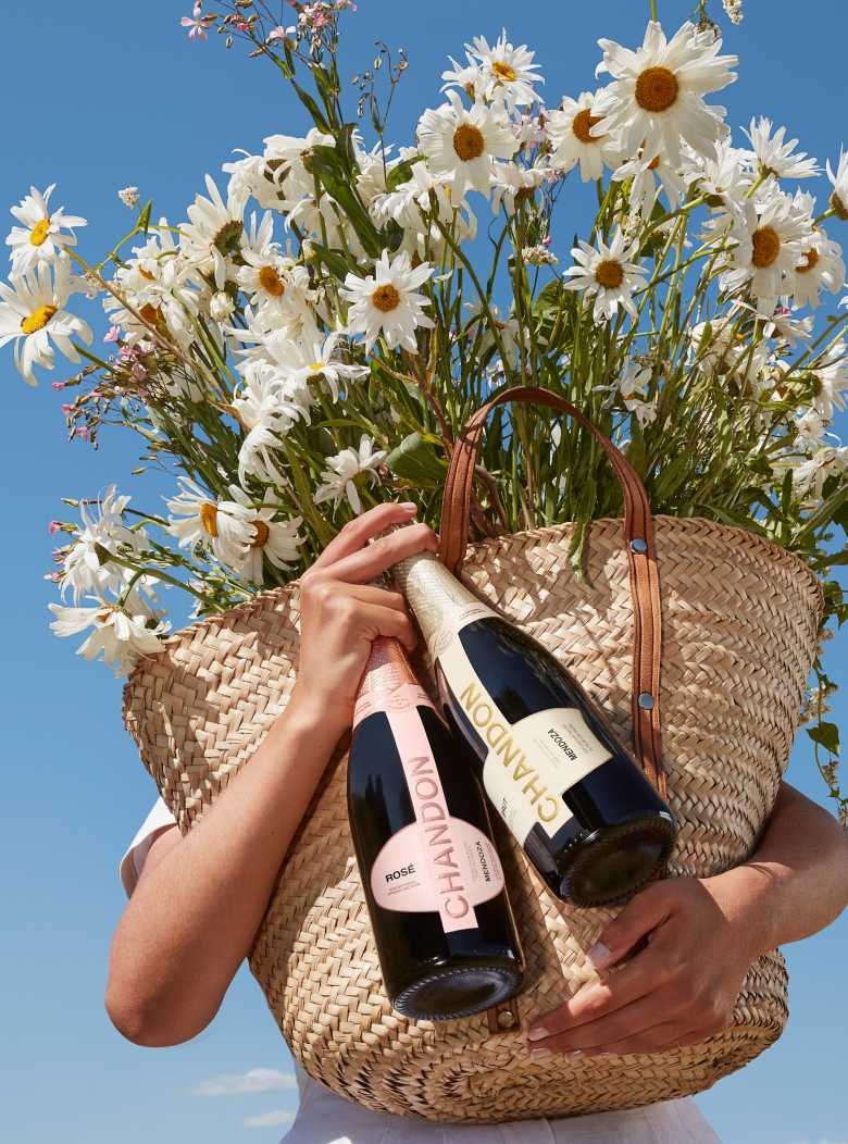 Persona sosteniendo un bolso con margaritas y dos botellas de espumante CHANDON Brut y Rosé Mendoza bajo un cielo azul.