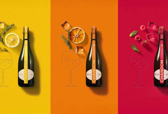 Trois bouteilles de Chandon Spritz, chacune avec une étiquette distincte (jaune pour Lemon & Verbena, rouge pour Berries et Hibiscus, orange pour Orange Peels & Spices). Chaque bouteille est accompagnée d'un verre stylisé gravé dans la surface et d'ingrédients frais assortis (citron, romarin et glaçons pour la jaune ; orange séchée, romarin et glaçons pour l'orange ; framboises, feuilles de menthe et glaçons pour la rouge), le tout présenté sur des fonds de couleurs vives coordonnées (jaune, orange, rouge).