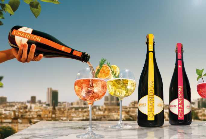 Drei Flaschen Chandon Spritz (Lemon & Verbena, Berries & Hibiscus und Orange Peels & Spices) werden neben den drei dazugehörigen Chandon Cocktails in Gläsern präsentiert, garniert mit Zitrone, Orange, Beeren, Rosmarin und Minze – vor einer sonnigen urbanen Außenkulisse. Zeigt die gesamte Bandbreite der erfrischenden Chandon Spritz Sorten, perfekt für sommerliche Zusammenkünfte und Feiern.
