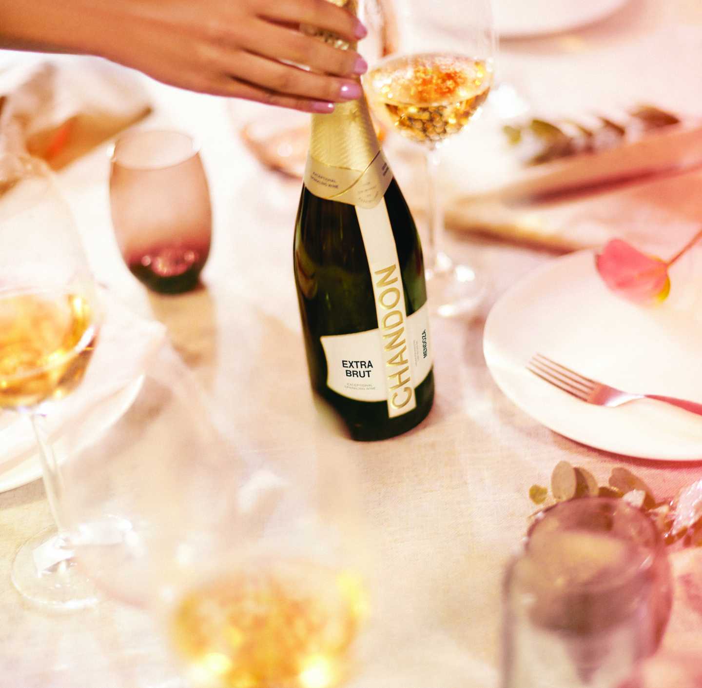 Mano femenina descorchando una botella de CHANDON Extra Brut en una mesa elegante con copas de vino.