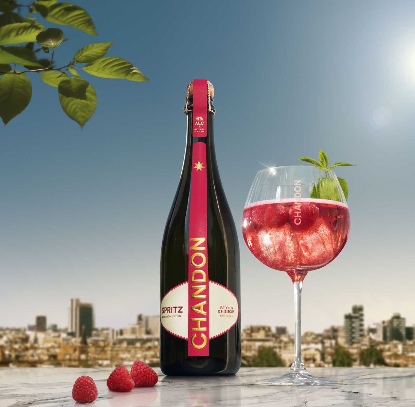 A bottle of Chandon Spritz Berries & Hibiscus next to a glass of Chandon Spritz cocktail garnished with fresh raspberries and mint leaves, set against a bright cityscape background under a clear sky.