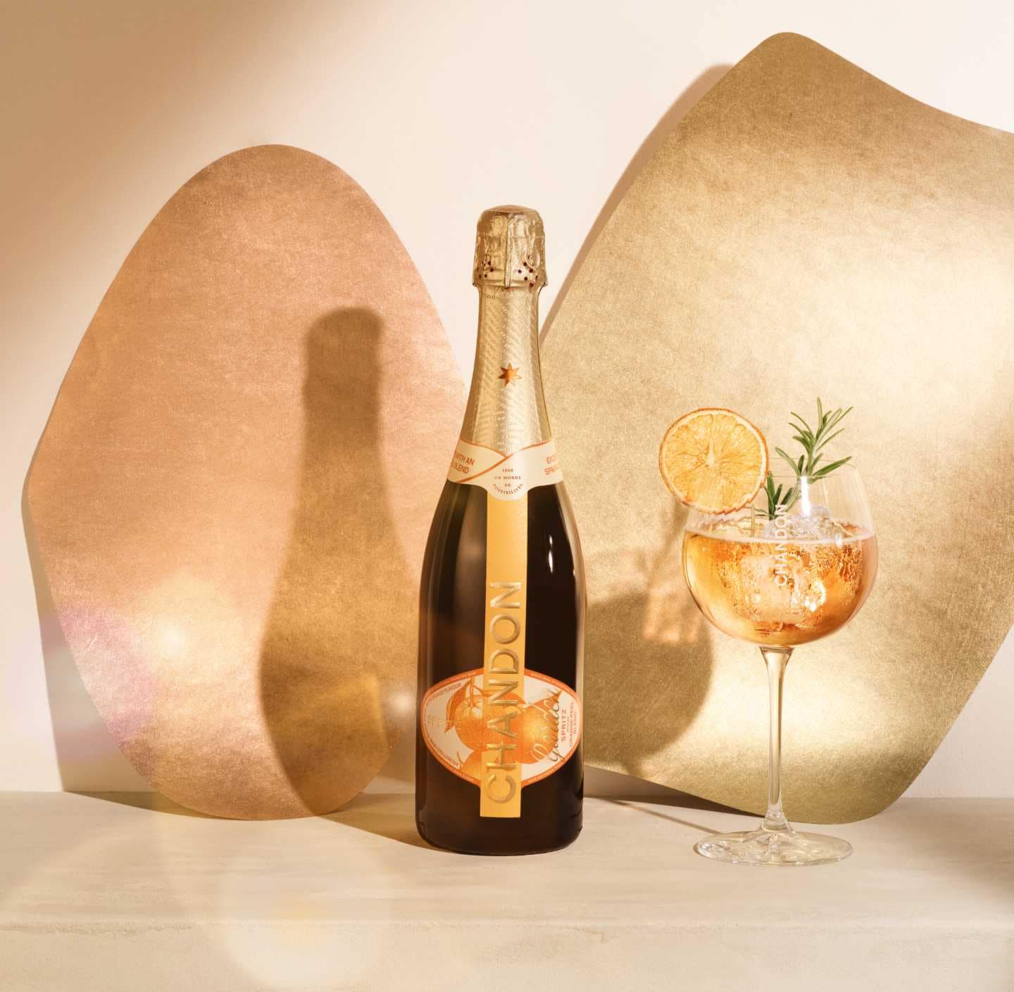 A bottle of CHANDON Garden Spritz and a glass of the spritz cocktail garnished with a dried orange slice and rosemary, against a modern, minimalist background of two abstract, textured panels in rose gold and gold hues.