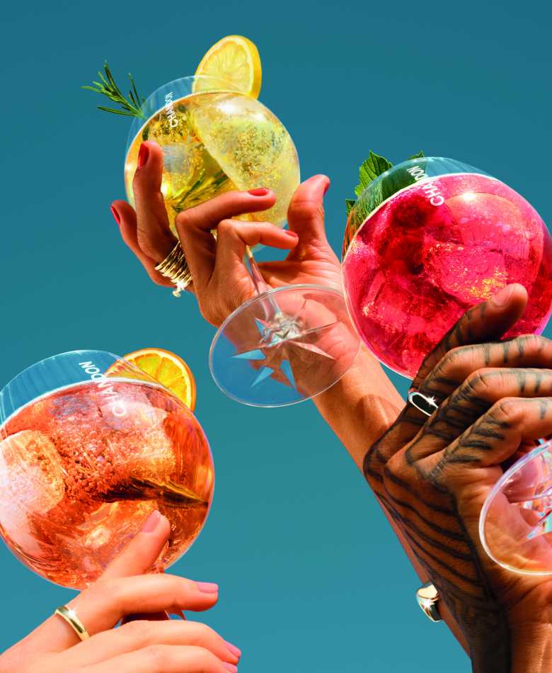 Close-up of three hands holding Chandon Spritz Orange, Lemon and Berries cocktail glasses, garnished with citrus and rosemary, against a vibrant blue sky