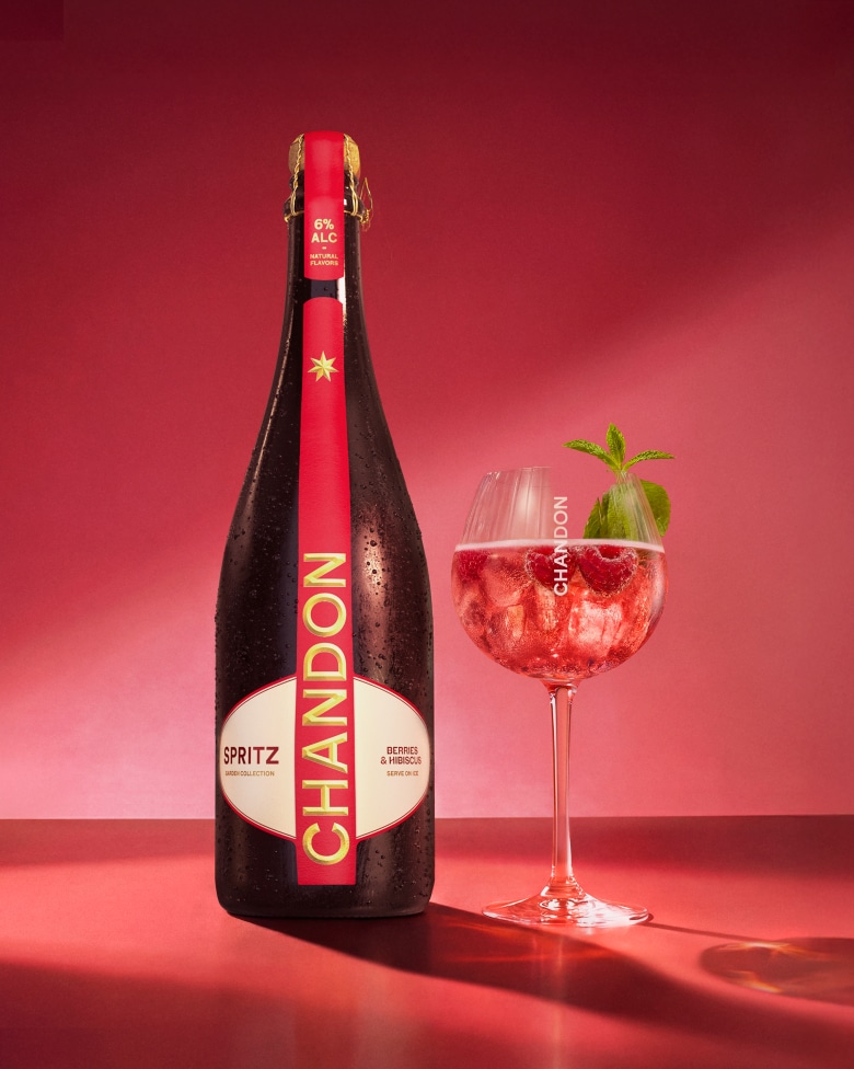 A bottle of Chandon Spritz Berries & Hibiscus next to an outline of a wine glass, with ice cubes, raspberries, and mint leaves scattered around on a bright red background