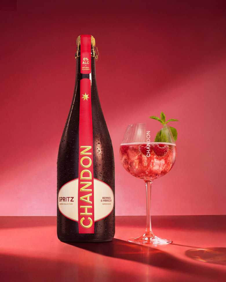 Eine Flasche Chandon Spritz Berries & Hibiscus neben einem Glas Chandon Spritz Cocktail, garniert mit frischen Himbeeren und einem Minzblatt, vor einem leuchtend roten Hintergrund.