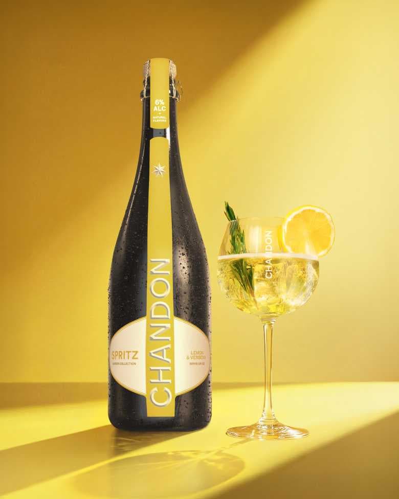 Eine Flasche Chandon Spritz Lemon & Verbena steht neben einem hohen Glas Chandon-Spritz-Cocktail, garniert mit einer Zitronenscheibe und einem Zweig Rosmarin, vor einem gelben Hintergrund mit Farbverlauf.