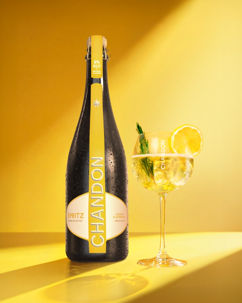 A bottle of Chandon Spritz Lemon & Verbena next to an outline of a wine glass, with ice cubes, a lemon slice, and a sprig of rosemary scattered around on a bright yellow background.