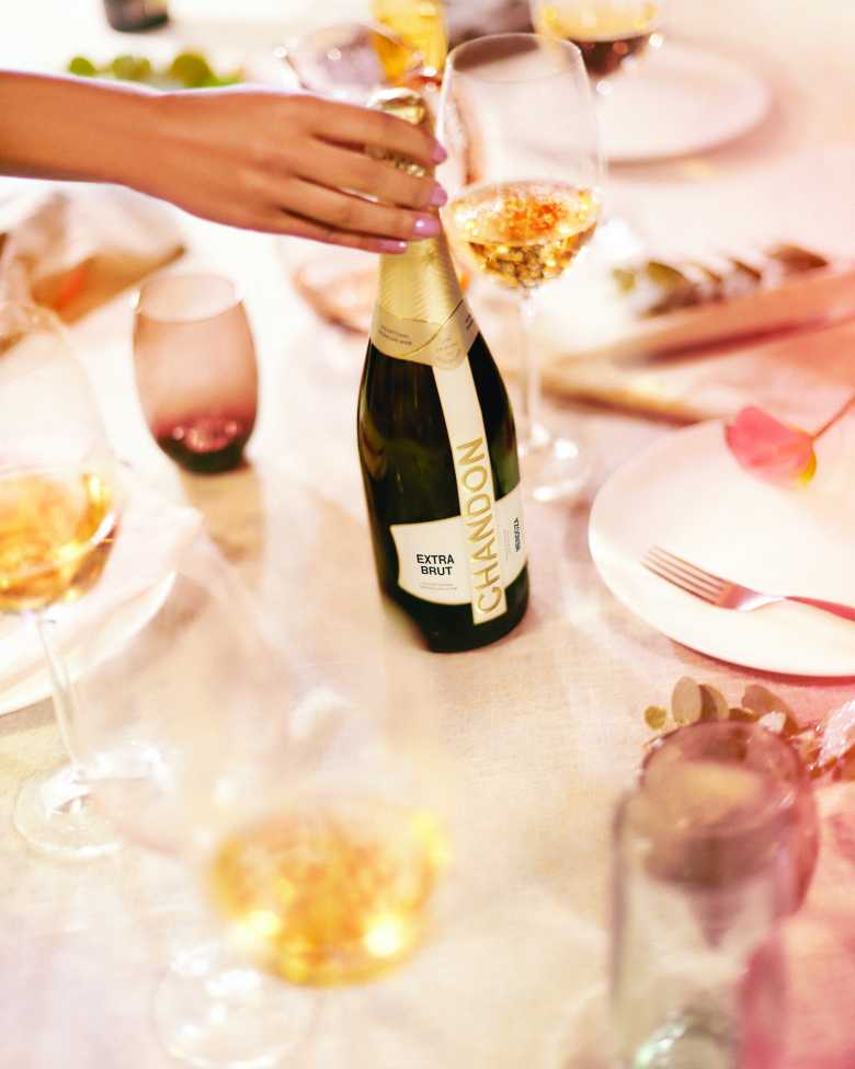Mano femenina descorchando una botella de CHANDON Extra Brut en una mesa elegante con copas de vino.