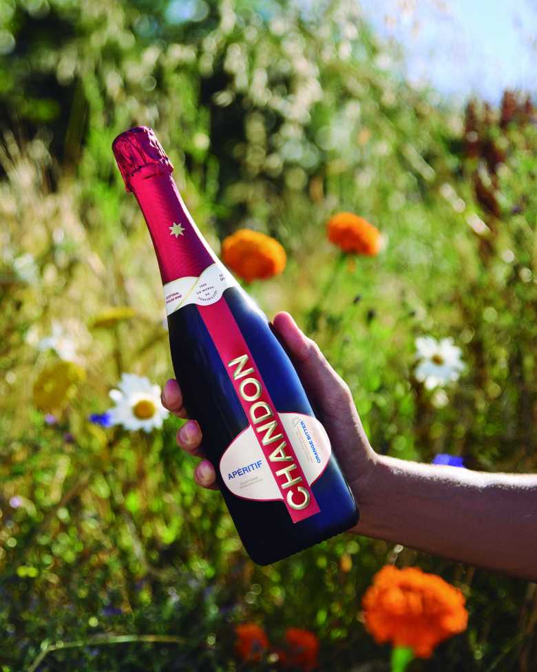 Botella de CHANDON Aperitif sostenida por una mano en un jardín de flores al aire libre.