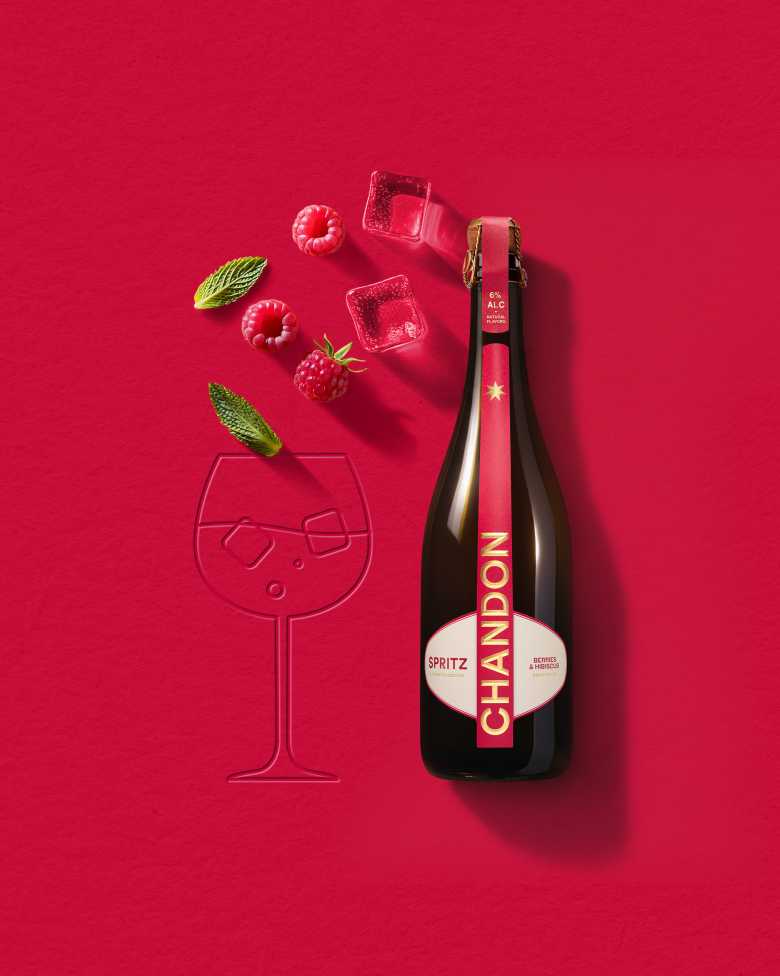 A bottle of Chandon Spritz Berries & Hibiscus next to an outline of a wine glass, with ice cubes, raspberries, and mint leaves scattered around on a bright red background