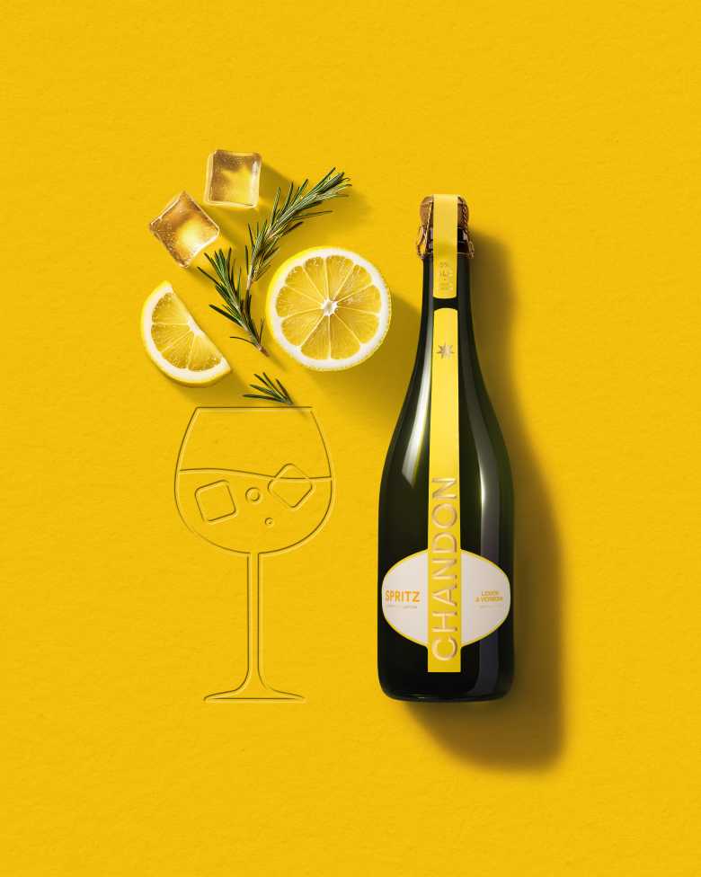 A bottle of Chandon Spritz Lemon & Verbena next to an outline of a wine glass, with ice cubes, a lemon slice, and a sprig of rosemary scattered around on a bright yellow background.