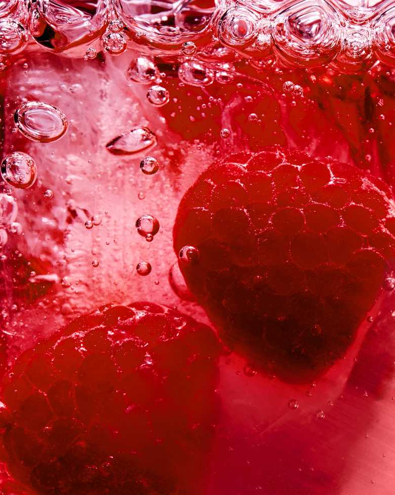 Gros plan sur deux framboises fraîches immergées dans un liquide rouge pétillant.