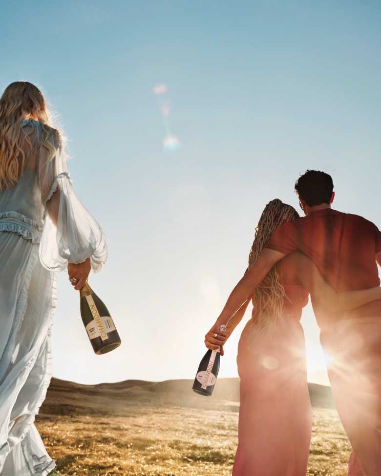 Amigos caminando juntos al atardecer por los viñedos de Mendoza sosteniendo botellas de Chandon Extra Brut y Rosé.