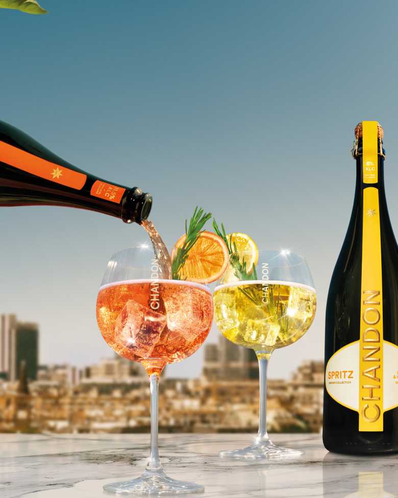 Drei Flaschen Chandon Spritz (Lemon & Verbena, Berries & Hibiscus und Orange Peels & Spices) werden neben den drei dazugehörigen Chandon Cocktails in Gläsern präsentiert, garniert mit Zitrone, Orange, Beeren, Rosmarin und Minze – vor einer sonnigen urbanen Außenkulisse. Zeigt die gesamte Bandbreite der erfrischenden Chandon Spritz Sorten, perfekt für sommerliche Zusammenkünfte und Feiern.
