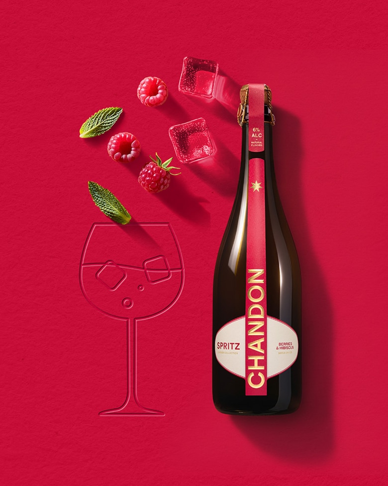  Eine Flasche Chandon Spritz Lemon & Verbena neben den Konturen eines Weinglases, mit Eiswürfeln, einer Zitronenscheibe und einem Rosmarinzweig, die auf einem leuchtend gelben Hintergrund verstreut sind.