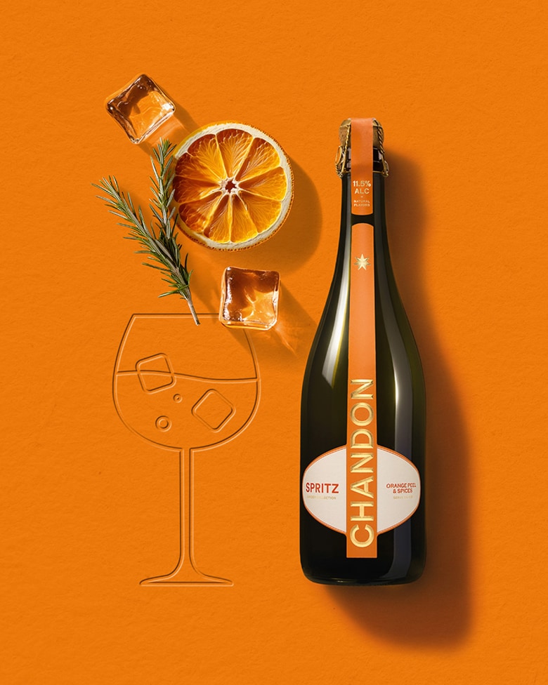 Chandon Spritz Flasche, getrocknete Orangenscheibe, Rosmarinzweig und Eiswürfel, arrangiert auf einem orangefarbenen Hintergrund neben einer geprägten Umrisszeichnung eines Weinglases.