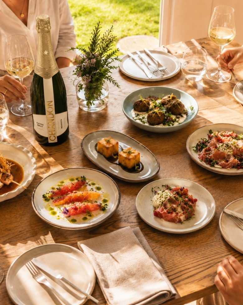 Botella de CHANDON Extra Brut junto a copas y platos gourmet en una mesa compartida.