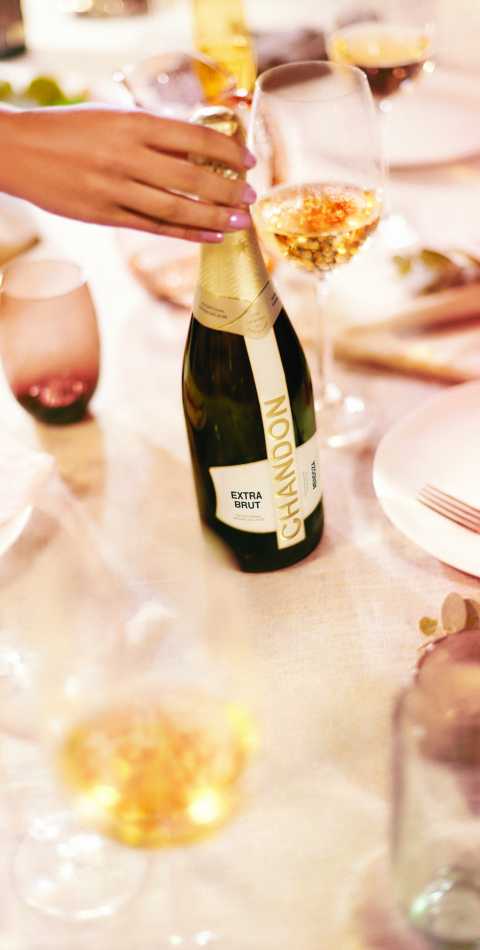 Mano femenina descorchando una botella de CHANDON Extra Brut en una mesa elegante con copas de vino.