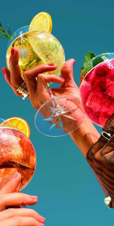 Three hands toasting glasses of Chandon Spritz cocktails: one with orange and rosemary, one with lemon and rosemary, and one with a red berry garnish, set against a bright blue sky.